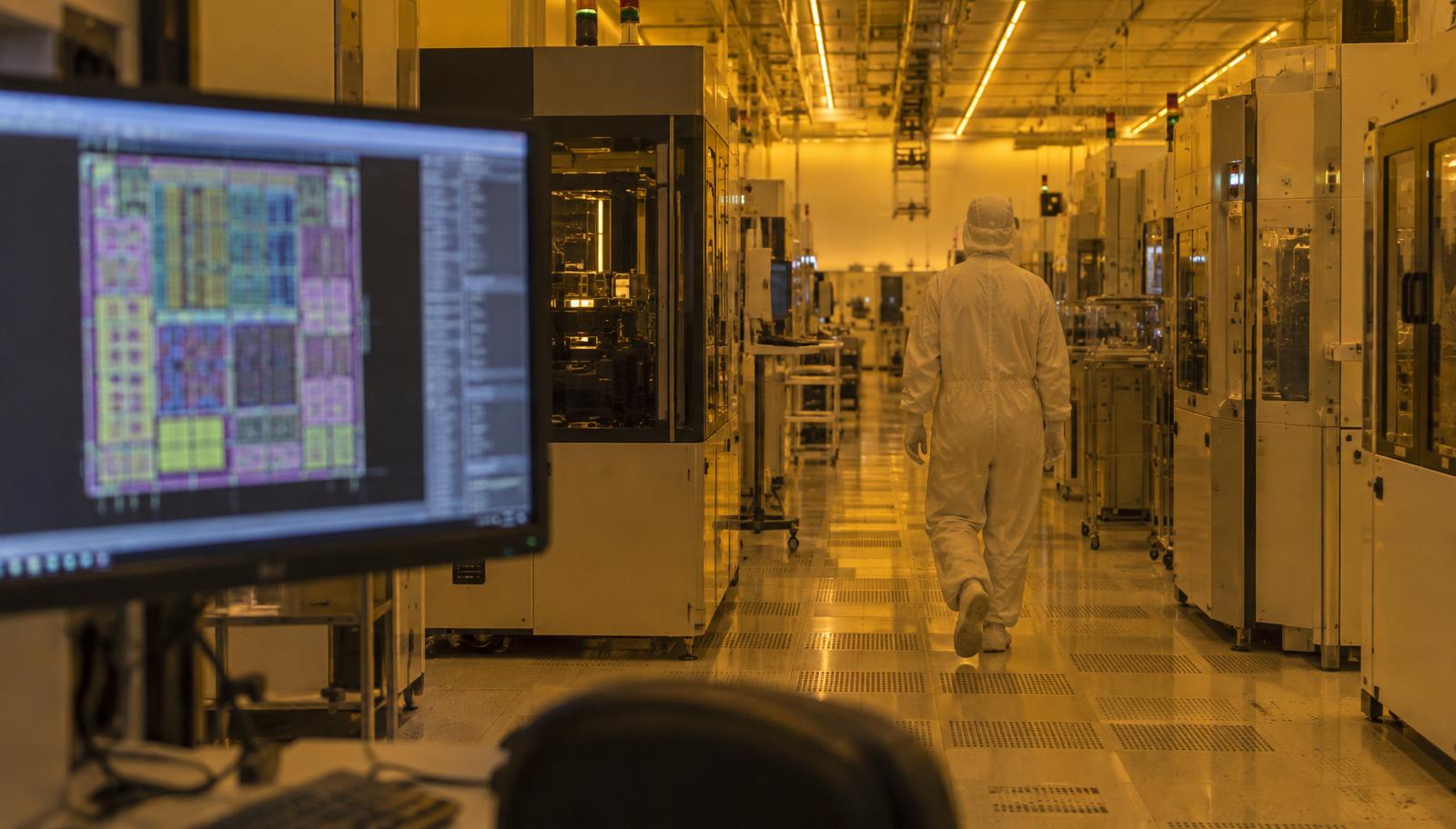 Une salle blanche sous la lumière ambrée de la lithographie, un technicien en combinaison blanche entre deux rangées d’équipements, un dessin de puce allumé sur un écran de conception au premier plan