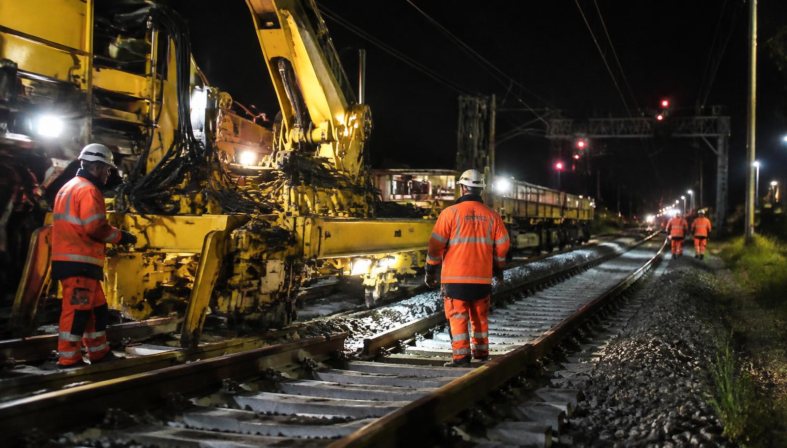 Rail workers in orange high-visibility gear walk beside a yellow track-renewal machine laying new sleepers and ballast at night under floodlights, with a signal gantry showing red lights further down the line