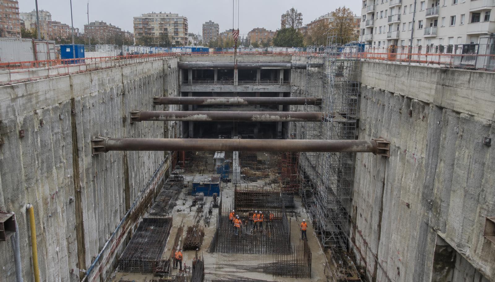 Looking down into a deep metro station excavation between apartment blocks, its concrete walls braced by steel struts, with construction workers in orange high-visibility gear standing on the reinforcement at the bottom